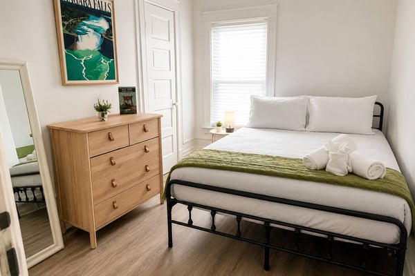 Bright bedroom with queen bed, wooden dresser, Niagara Falls poster, and large window with white trim